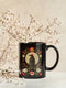 A black ceramic mug featuring a gothic design of a raven against a golden full moon, framed by a floral wreath of coral-colored daisies, white chrysanthemums, and delicate botanical elements. The design includes rich green foliage and small red buds arranged in a symmetrical pattern. The mug has a classic C-shaped handle with a glossy finish.